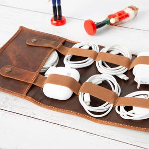May include: A brown leather cable organiser with multiple compartments, holding white cables, chargers, and a pair of white earbuds. The organiser is secured with brown elastic straps and features a button closure. A small toy soldier and a nutcracker are in the background.