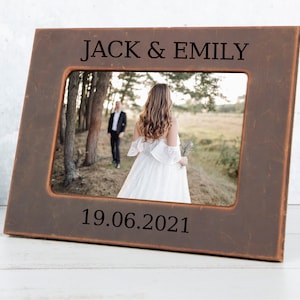 May include: A brown wooden picture frame with a black engraved inscription that reads "JACK & EMILY 19.06.2021". The frame holds a photo of a couple in a field.