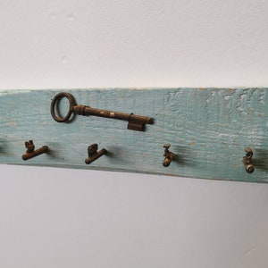 Könnte beinhalten: Ein rustikaler Schlüsselhalter aus Holz mit einer abgewetzten hellblauen Lackierung. Er verfügt über fünf antike Schlüsselhaken und einen dekorativen antiken Schlüssel.
