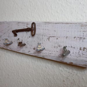 Könnte beinhalten: Ein rustikaler, weiß lackierter Schlüsselhalter aus Holz mit einem Used-Look. Er verfügt über einen großen Schlüssel im antiken Stil und mehrere kleine Metallhaken zum Aufhängen von Gegenständen. Der Halter ist an einer strukturierten Wand befestigt.
