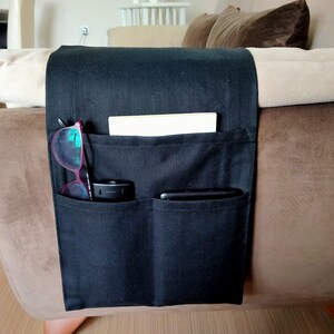 May include: Black fabric armrest organiser with multiple pockets. It has a book, glasses, and two phones in the pockets.