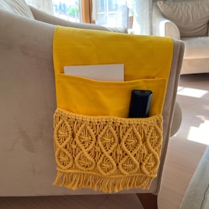 May include: A mustard yellow macrame armchair tidy with pockets, holding a remote control and papers. The caddy is attached to the arm of a beige armchair. The macrame design adds a decorative touch.