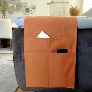 May include: A brown fabric couch armrest organizer with three pockets. The organizer has a white piece of paper in the top pocket and a black phone in the middle pocket.