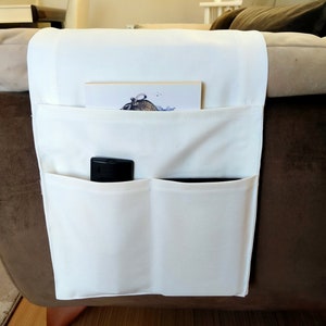 May include: White fabric organiser with multiple pockets attached to the arm of a brown sofa. The organiser has a remote control and a book inside.