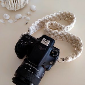Op de afbeelding: Een zwarte camera met een witte gehaakte cameraband. De camera heeft een lens met de tekst "DT 18-55mm F3.5-5.6 SAM" en "Sony" erop gedrukt.