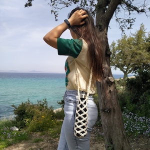 May include: A person wearing a green and white shirt and blue jeans with a white rope bottle holder. The person is standing next to a tree with a view of the ocean in the background.