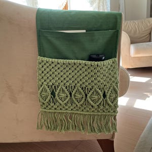 Può includere: Un organizer per bracciolo del divano in macramè verde con due tasche e frange. L'organizer è attaccato a un divano beige. Un telecomando è visibile nella tasca inferiore. Il design in macramè presenta forme a diamante.