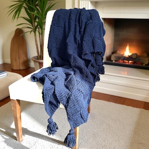Puede incluir: Una manta de punto azul marino con un patrón geométrico está sobre una silla blanca. La manta tiene borlas en las esquinas y se muestra frente a una chimenea con fuego. La habitación tiene una paleta de colores neutros.
