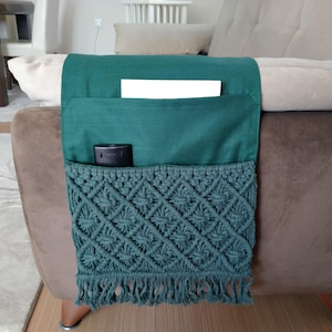 May include: A teal blue couch armrest organiser with a crocheted pocket and a fringe. The organiser has a pocket for a remote control and a small compartment for other items.
