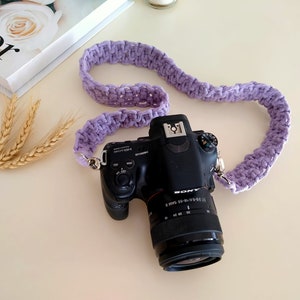 Puede incluir: Una cámara Sony negra con una correa de cámara de crochet morado. La cámara tiene un objetivo con un anillo plateado.