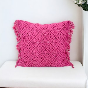 May include: A pink macrame pillow with a diamond pattern and fringe trim. The pillow is sitting on a white bench.