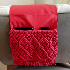 May include: Red crocheted couch arm organiser with fringe. The organiser has two pockets, one for a phone and one for a remote control.