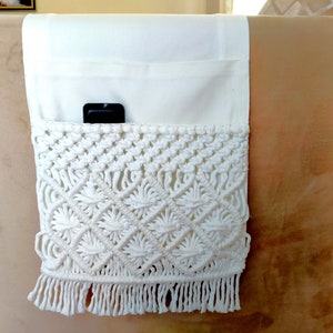 May include: White crocheted armrest organiser with a pocket and fringe. The organiser is attached to the armrest of a sofa.