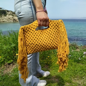 May include: A yellow crocheted bag with long fringe tassels. The bag is held by a person's hand in front of a blue body of water.
