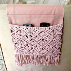 May include: A pink macrame couch arm organizer with fringe. Two black remotes are inside the organizer.