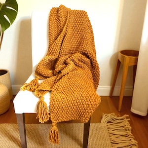 May include: Mustard yellow knitted throw blanket with tassel accents draped over a white chair. The blanket has a textured, raised stitch pattern. The tassels are a matching mustard yellow color. Home decor item.