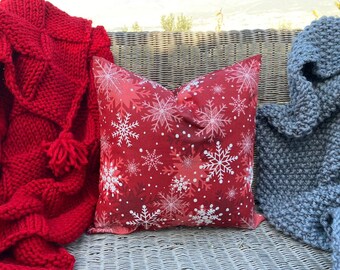 May include: A red and white throw pillow with a snowflake pattern. The pillow is on a wicker couch with a red knitted blanket and a blue knitted blanket.