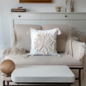 May include: A white couch with a white throw pillow featuring a macrame design. The couch is in a living room with a wooden coffee table and a rug.