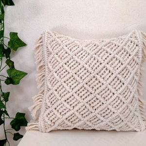 Könnte beinhalten: Ein cremefarbenes Makramee-Kissen mit einem Rautenmuster und Fransenbesatz. Das Kissen ist aus Baumwollschnur gefertigt und eine großartige Ergänzung für jedes Boho- oder moderne Wohndesign.