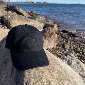 May include: A black baseball cap with an embroidered outline of the Great Lakes on the front. The cap is sitting on a large rock near a body of water.