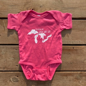 Puede incluir: Un body de bebé rosa con un contorno blanco de los Grandes Lagos y el texto "I'm living the Great Lake Life".