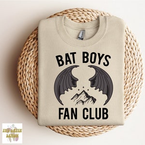 Puede incluir: Una sudadera beige con texto negro que dice "Bat Boys Fan Club". El texto está rodeado por un gráfico de dos alas de murciélago negras con una cordillera de montañas entre ellas.