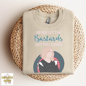 May include: Beige sweatshirt with the text "Never Let The Bastards Get You Down" in pink and blue lettering. A cartoon illustration of a woman with blonde hair is in the center of the sweatshirt.