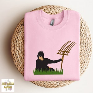 May include: A pink sweatshirt with a graphic of a person in sunglasses holding a pitchfork in a field of green grass.