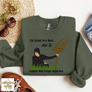 May include: A green sweatshirt with a black and white illustration of a person holding a pitchfork. The text on the sweatshirt reads "I'm Trying Very Hard Not To Connect With People Right Now".