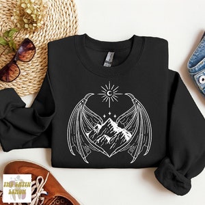 Puede incluir: Sudadera negra con un diseño gráfico blanco de una cordillera con alas y un sol y una luna encima.