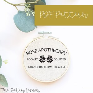 Può includere: Un telaio da ricamo con il testo "Rose Apothecary", "Locally Sourced, Handcrafted with Care" e due illustrazioni di rose nere. Le parole "PDF Pattern" sono in un rettangolo dorato. Sfondo bianco.