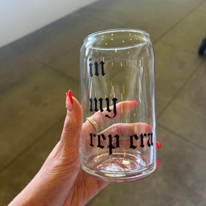 Puede incluir: Un vaso transparente en forma de lata con las palabras "in my rep era" impresas en negro con fuente gótica. El vaso está sostenido por una mano con esmalte de uñas rojo.