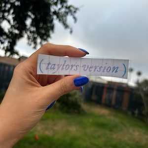 Könnte beinhalten: Ein rechteckiger Aufkleber mit dem Text "(taylor's version)" in einem hellvioletten Farbverlauf. Der Aufkleber wird vor einem bewölkten Himmel und grünem Laub gehalten.