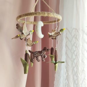 May include: A hanging mobile featuring felt figures suspended from a circular jute-wrapped frame. The elements include two flying ducks with yellow beaks, a white crescent moon, a brown dog with white spots, and two brown cattail plants.