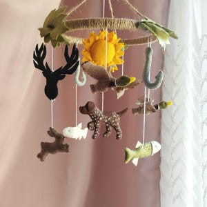 May include: A hanging nursery mobile featuring felt cutouts of a black deer head, a yellow sun, a brown dog, a brown duck, a white fish, a green fish, and two grey fishing hooks suspended from a circular frame wrapped in natural jute twine.