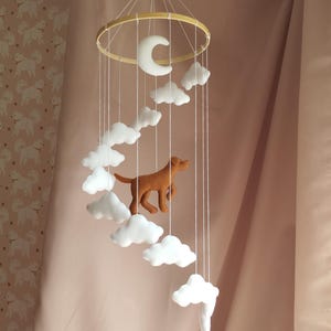May include: A hanging mobile featuring a circular wooden frame with white strings suspending a brown felt dog, a white felt crescent moon, and multiple white felt clouds arranged in a spiral pattern, creating a layered and dimensional display.