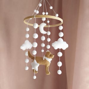 May include: A baby mobile featuring a wooden hoop with hanging white felt clouds, moon, stars, and pom-poms. A brown felt dog with a white scarf is suspended below. The mobile is designed for a nursery.