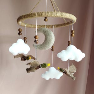 May include: A handmade baby mobile featuring a jute-wrapped ring, wooden beads, and felt elements. The mobile includes a gray felt moon, white felt clouds, and brown felt ducks with green and yellow accents. The mobile hangs from white string.