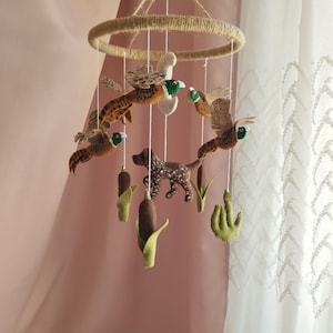 May include: A hanging mobile featuring felt figures of brown and green pheasants in flight, a brown spotted dog, and green cattail plants suspended from a circular frame wrapped in tan twine with white strings attached to each individual element.