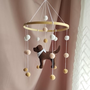 May include: A hanging mobile featuring a circular wooden frame suspended by white strings. A brown felt dog figure with a white collar hangs in the center, surrounded by multiple white, beige, and mustard yellow pom-poms attached to the strings.