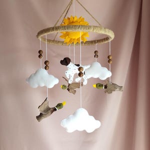 May include: A hanging mobile featuring a circular jute-wrapped frame with a central yellow felt sun. Suspended from the frame are white felt clouds, brown and white spotted dog figures, and brown ducks with yellow beaks, accented by wooden beads.