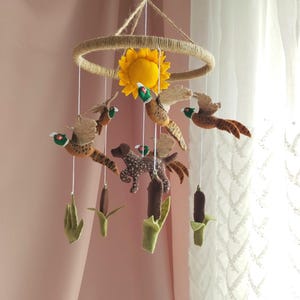 May include: A hanging nursery mobile featuring a circular jute frame with a central yellow felt sunflower. Suspended from the frame are felt figures of brown and green pheasants in flight, a brown spotted dog, and green cattail plants on white strings.