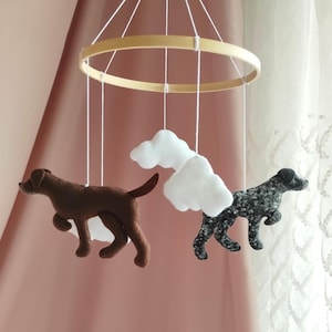 May include: A hanging mobile featuring a circular wooden frame with white strings suspending two felt dog figures and white felt clouds. One dog is solid brown, and the other is black with a white speckled pattern, both in a running pose.