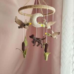 May include: A handmade baby mobile featuring felt ducks, a crescent moon, a dog, and cattails. The ducks have brown wings and yellow beaks. The dog is brown with white spots. The mobile is suspended from a jute-wrapped ring.