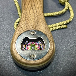 May include: A wooden bottle opener with a metal cap opener and a colorful mandala design in the center. The opener has a green paracord lanyard with a brass bullet accent.