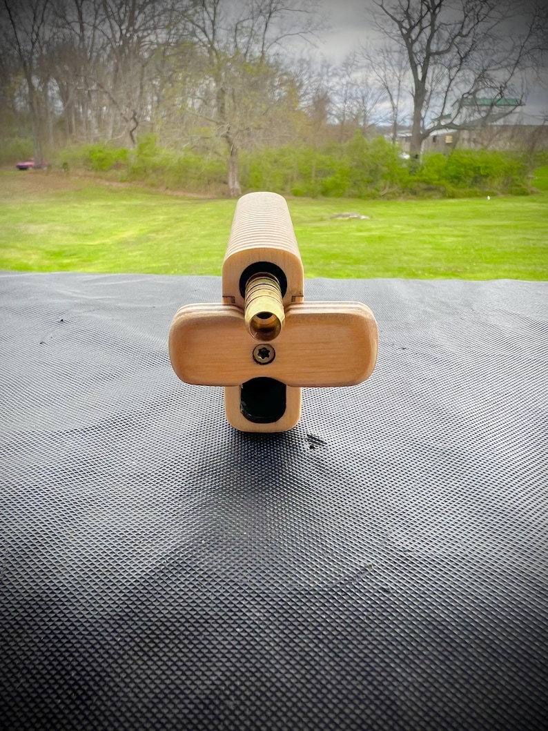 Puede incluir: Un modelo de arma de madera con un ca&ntilde;&oacute;n de lat&oacute;n y un dise&ntilde;o en forma de cruz. El arma est&aacute; apoyada sobre una superficie negra texturizada.