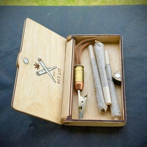 Puede incluir: Caja de madera con tapa con bisagras, grabada con una hoja de cannabis y porros cruzados, y el texto "GET LIT 420". En el interior hay porros pre-enrollados, un clip met&aacute;lico y un soporte con forma de bala.