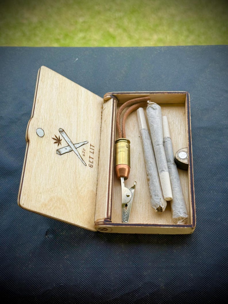 Puede incluir: Caja de madera con tapa con bisagras, grabada con una hoja de cannabis y porros cruzados, y el texto "GET LIT 420". En el interior hay porros pre-enrollados, un clip met&aacute;lico y un soporte con forma de bala.