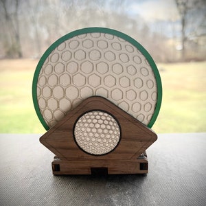 Puede incluir: Un juego de posavasos de madera con forma de bola de golf con un posavasos con borde verde y un soporte. El posavasos tiene una superficie texturizada que se asemeja a una bola de golf.
