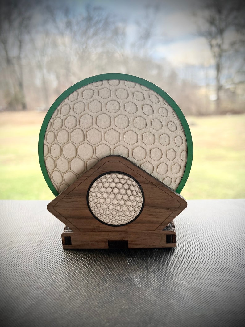 Puede incluir: Un juego de posavasos de madera con forma de bola de golf con un posavasos con borde verde y un soporte. El posavasos tiene una superficie texturizada que se asemeja a una bola de golf.
